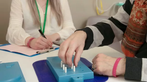 A man with grey hair, wearing a scarf and jumper is sat at a table doing a test involving putting white pegs in holes in a blue tray. A woman with blonde hair wearing a lanyard is taking notes. 