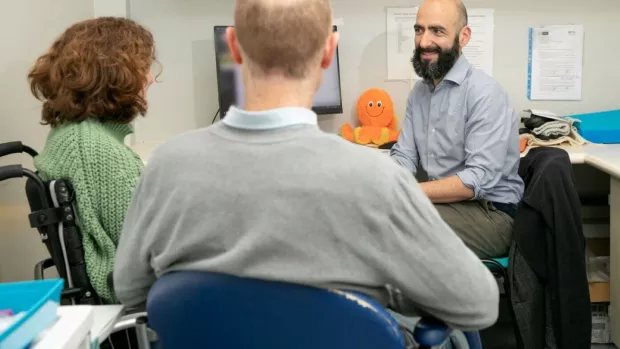 Three people sit in a hospital consulting room. A trial participant is sitting in a wheelchair. Her husband has his back to the camera. A researcher with a beard is smiling at the. An orange fluffy octopus is on a desk behind him. 