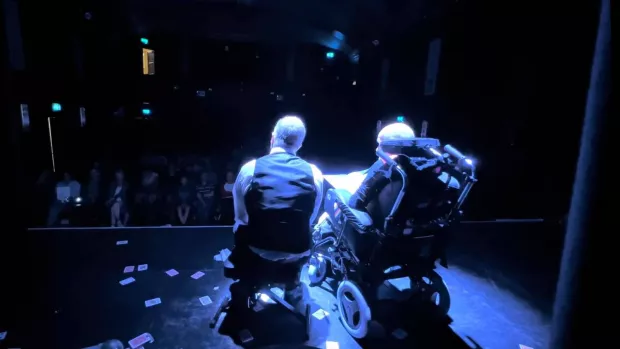 A figure in a wheelchair and a crouching figure in a waistcoat are on a dark stage looking out into an audience, they're surrounded by playing cards scattered over the stage floor