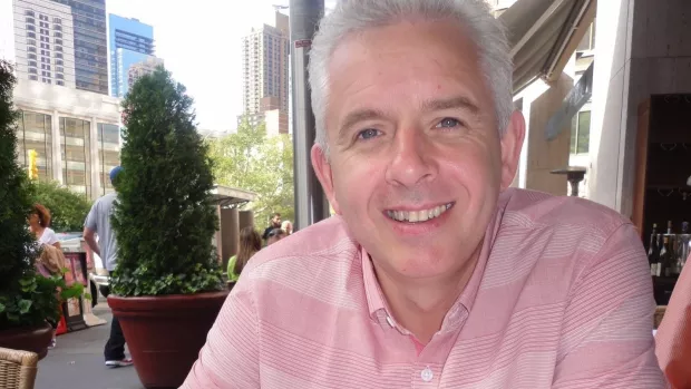 Head and shoulders of man with grey hair, wearing a pink shirt and smiling at the camera