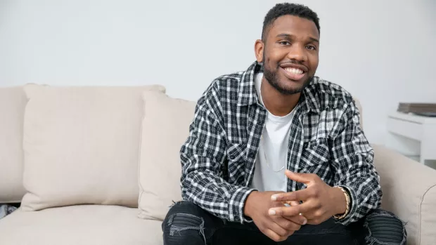 Jordan is sitting on a cream sofa wearing a dark blue, checkered shirt with a white t-shirt underneath. He's smiling at the camera.