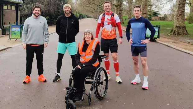 The photo shows Sara Weller in Battersea Park in her assisted wheelchair. Her training team stand behind her, smiling at the camera. 