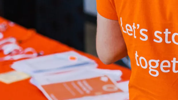 The back of an orange MS Society t-shirt which reads "Let's stop MS together", in the background an orange covered table is laid out with lanyards and leaflets
