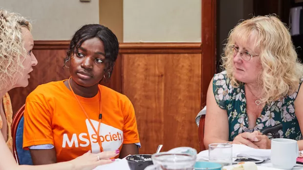 in the foreground there are two men in profile to the left and right. Between them at the opposite end of a table sit three women in conversation. The woman in the centre wears an MS Society orange t-shirt