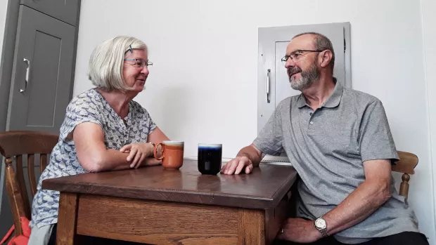 Brian and Diane Gist sit facing each other over a kitchen table, they're smiling and relaxed looking with mugs on the table between them. 