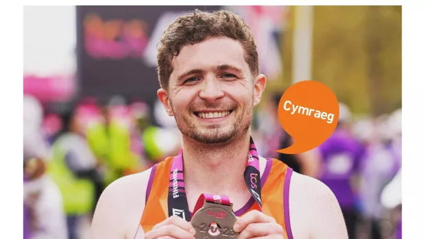 Image shows a man with short brown hair smiling. He is holding a medal after he completed the London Marathon 2023. Cymraeg logo. 