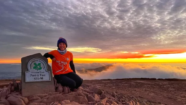 Image shows a woman smiling wearing an orange t shirt at the top of Pen y Fan