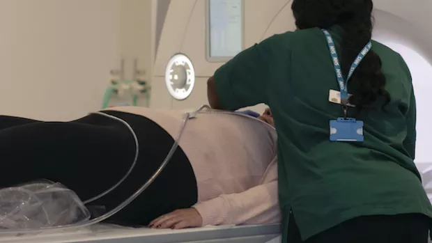 A woman in black trousers and pink top lies down about to have an MRI scan, a radiologists in a green top with long black hair is leaning over her
