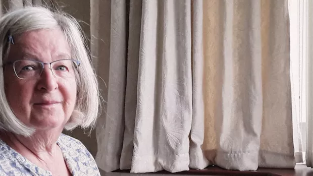 A woman with glasses and light bobbed hair sits in a dark chair in a sunny room with pale beige curtains, she looks at the camera with resolve.