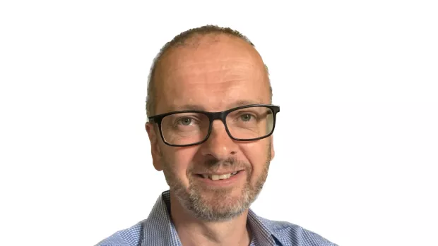A man with short grey hair and glasses in a blue and white checked shirt