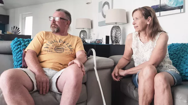 A man in shorts and a yellow t-shirt sits on a stone coloured sofa with a walking aid propped up against it, a woman in jean shorts and sleeveless top sits next to him. Both stare to the left as if out of a window. 