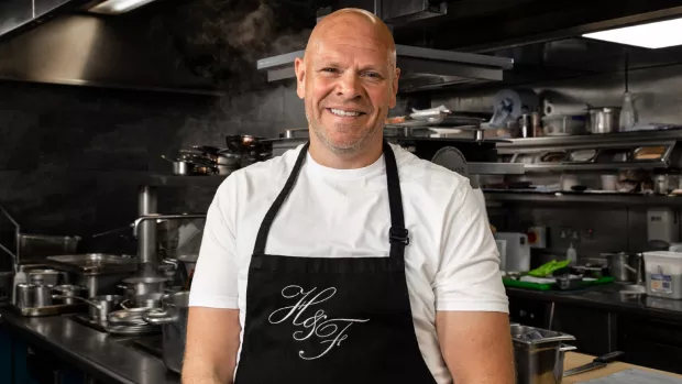 Tom is standing in his restaurant's kitchen wearing a white t-shirt and a black apron. He's smiling at the camera with his arms down by his sides.