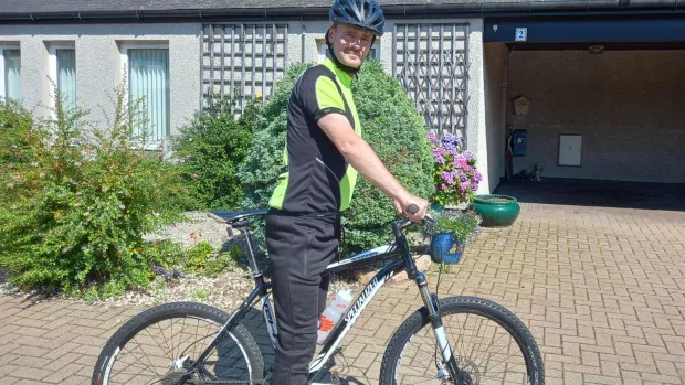 A man in cycling gear and helmet astride a mountain bike in front of a bungalow. 