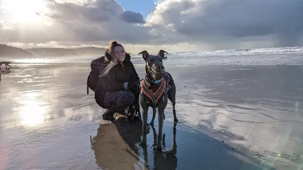 Amy Johnson kneels down on the wet shore of a beach with her greyhound, bleu. The horizon behind them is covered with white clouds with a bright sun breaking through them and reflecting on the wet shore