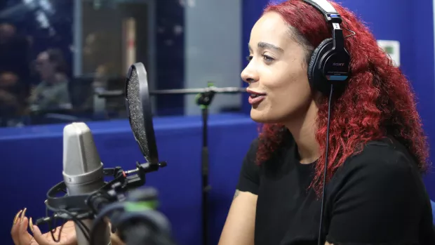 Roxy is sitting in a podcast recording studio with headphones on, talking into the microphone.