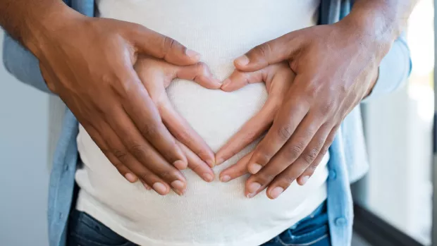 Man's hands over women's hands forming a heart over her pregnant belly. 