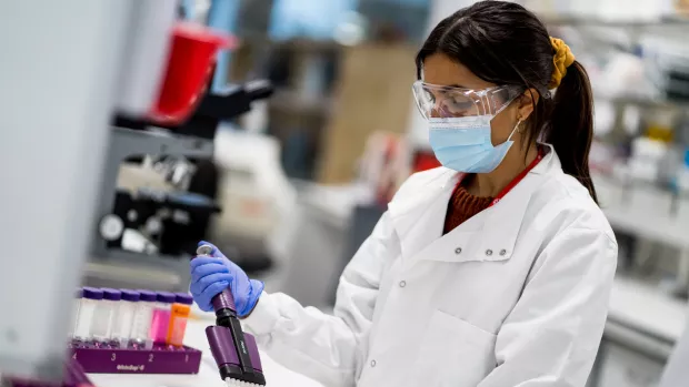 A researcher in a lab coat, protective goggles and mask