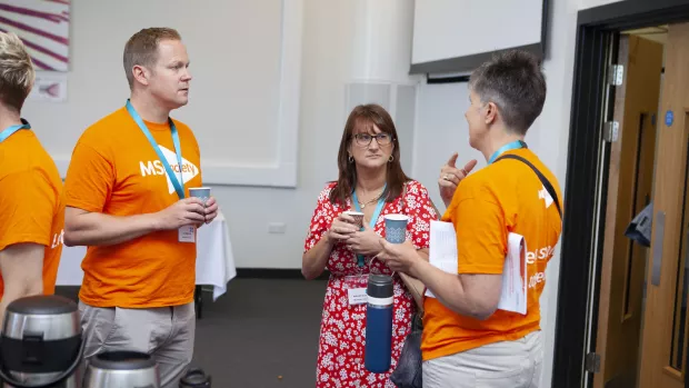 Three people stand in discussion, two wear orange MS Society t-shirts.