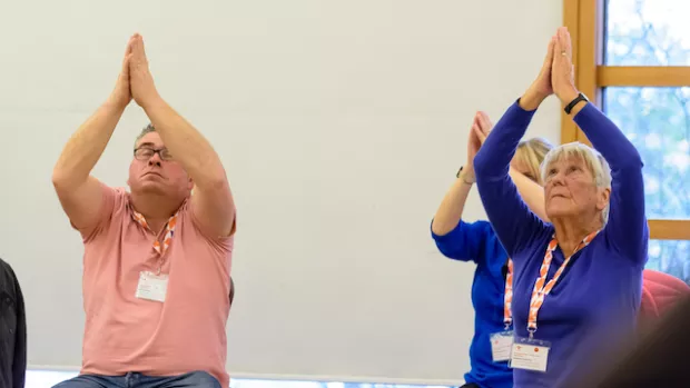 a group of people are doing seated yoga, with their palms held together and arms upraised