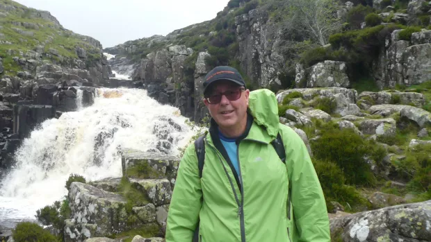 Gary wears a green waterproof jacket, sunglasses and cap and stands in front of a waterfall
