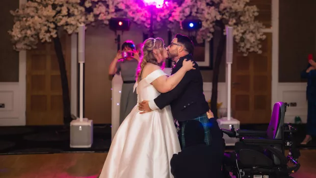 A wedding couple embrace for their firs dance, she is wearing a white wedding dress and he is in a kilt, behind him is his wheelchair from which he's just stood up