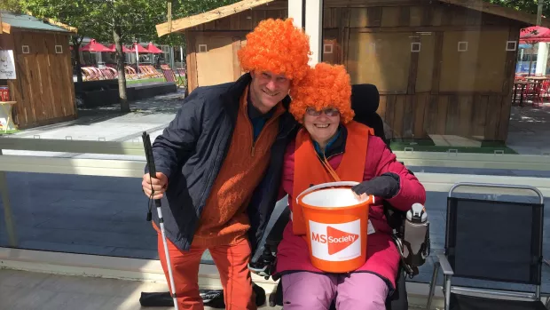 Jo is in her wheelchair holding an orange MS Society collection bucket and wearing an orange curly wid, to her right a man in similar wig crouches to pub his arm around her, they're both smiling with big smiles at the camera