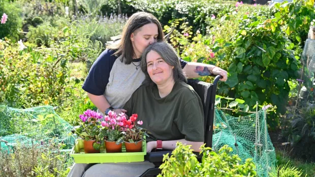 Sarah stands by her wife Karine, who is using a wheelchair. Sarah kisses her on the top of her head.
