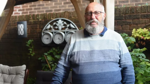 Mike has a thick white beard and wears glasses and a jumper with thick vertical stripes going from light to dark blue as they descend. Behind mike is a garden wall and some plants.