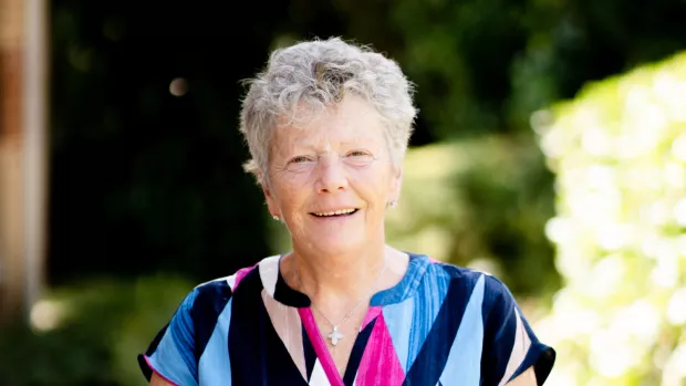 Siobhan has short wavy light grey hair and wears a colourful short sleeved top in pink and blue, behind them is the dappled light of of an out of focus summer garden.