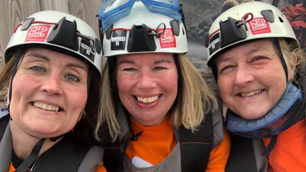 Three friends take a group selfie, they're wearing helmets with ZIP World stickers on them and orange MS Society t-shirts, they look excited. 