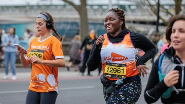 A person in an orange MS Society vest is running in a marathon flanked by two other runners