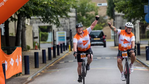 Two cyclists in orange MS Society cycling vests raise their fists in triumph as they cross a finish line
