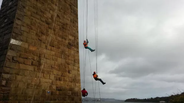 On the left is the side of a high brick tower or chimney with countryside far in the low distance. Three people in orange MS Society t-shirts abseil down the side of the building.