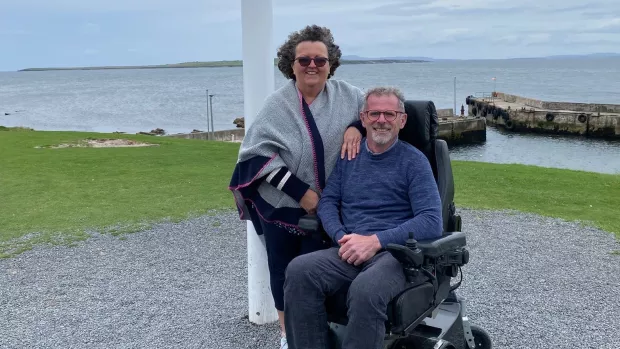 Marie stands by Stuart, who is using his wheelchair, in a waterside location.