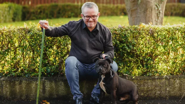 A man with a walking stick sits outside, with his dog