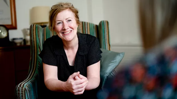 A person is sitting in a green stripy armchair. They are leaning forward with their elbows resting on their knees and holding their hands together. They are smiling and looking at another person that is out of focus.