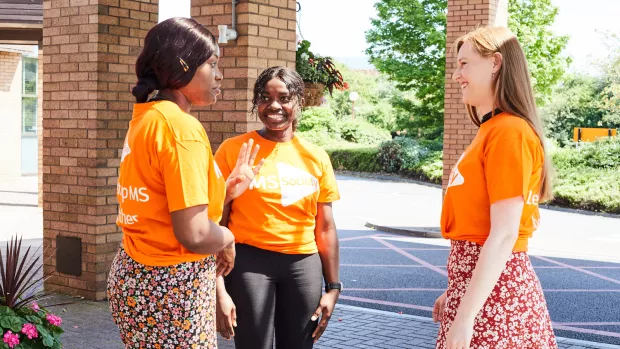 three people in MS Society t-shirts stand outside a building chatting and smiling