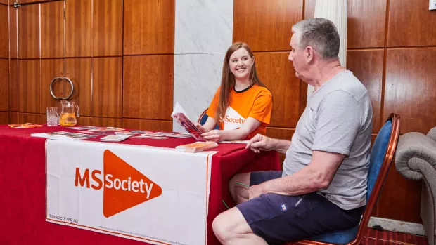 Two people sat at a table with an MS Society banner. One person is holding a booklet and smiling at the other person