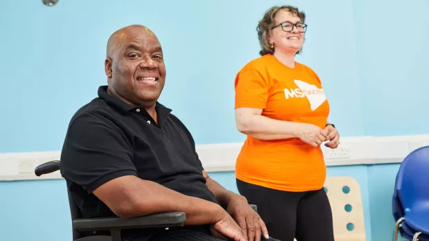 A man in a wheelchair smiles at the camera, with a person in an MS Society t-shirt in the background