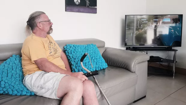 Ian Crosby is sat on a grey sofa with a walking aid looks at a television screen in the corner of a room.