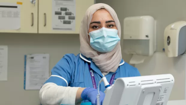 In a consultation room, a nurse in a hijab wears a face mask, gloves, and a lab coat while looking directly at the camera.