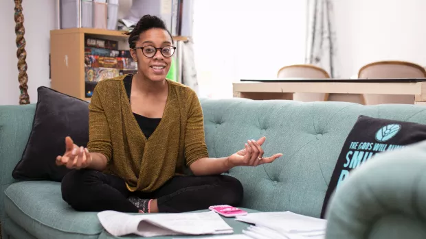 A woman with glasses sits on a sofa surrounded by documents