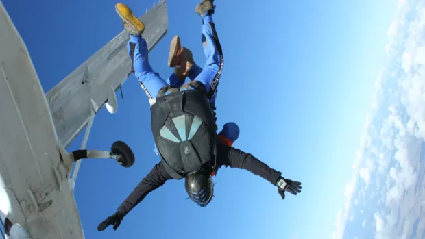 Two people strapped together skydive from a plane to the right there's a horizon of clouds in the distance far below and to the left there's the underside of the plane they've jumped from.