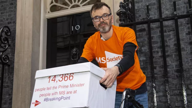 A man in an MS Society orange t-shirt is outside 10 Downing Street with a white archive box. The front of the box reads: "14,366 people tell the Prime Minister people with MS are at #BreakingPoint"