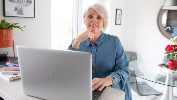A woman with light hair wears a denim coloured shirt and sits at an open laptop. She rests her chin on her hand and smiles casually at the camera