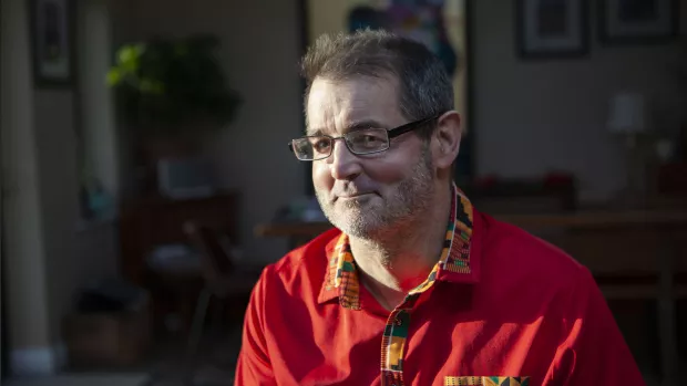 Mark sat in his living room wearing a red shirt, smiling slightly into the camera