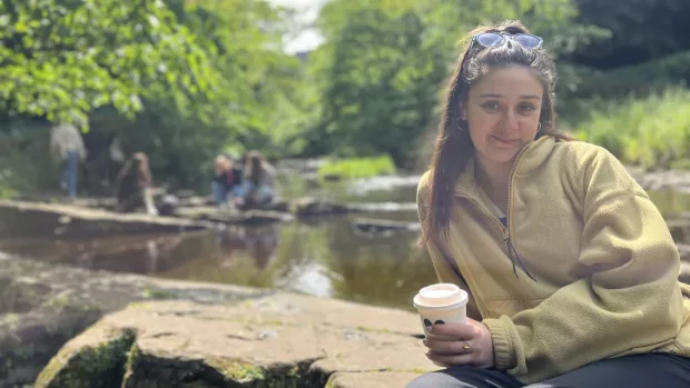 Yasmin reclines against some rocks in the sun, in front of a body of water, holding a coffee cup.