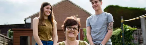 Photo shows a mum sitting in her garden, with her two teenage children standing behind her.