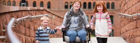 A photo of a mother in a wheelcahir with her two young childeren on a bridge