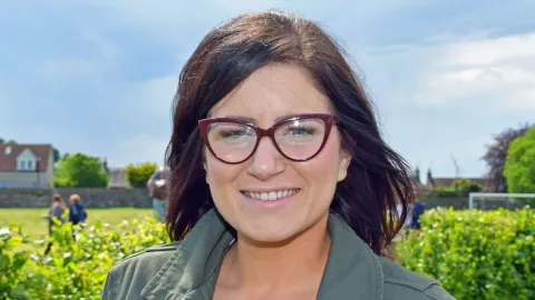 Headshot of Shirley smiling. She has long brown hair and glasses, and is standing outside by a field. 